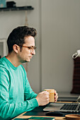 Side view of male distance worker in eyeglasses with mug of hot drink against netbook in house
