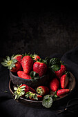 Appetizing fresh ripe juicy strawberries with green leaves served in bowl on dark wooden table with black background