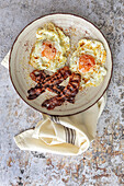 Overhead view of tasty sunny side up eggs with fried bacon strips on plate above towel