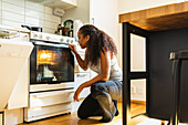 Young African American housekeeper with curly hair wearing casual clothes squatting and looking inside of oven while observing cooking process