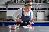 Woman in apron by placing strawberries cut from a bowl in the bowl with tongs in delicious dessert in kitchen of modern restaurant