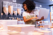 Young African American woman in uniform and glasses with curly hair checking goods in glass boxes and using laptop while working in zero waste grocery store