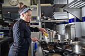 Side view of woman in dark uniform pouring oil on hot frying pan while working in professional kitchen of restaurant