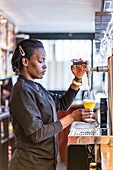 Side view of black female barkeeper pouring cold beer in glass in bar