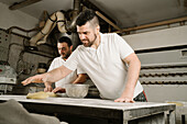 From below mature bearded ethnic bakers forming bread from dough at table with flour and bowl in bakery