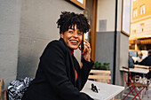 Excited curly haired African American female in trendy outfit sitting at table in outdoor cafe having phone conversation looking away