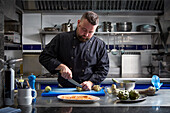 Bearded male chef cutting fresh artichokes while preparing delectable dish on table in kitchen of contemporary restaurant