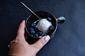 From above cropped unrecognizable person hand holding bowl full of delicious ripe blueberries placed on black background