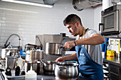 Concentrated young ethnic male cook with dark hair in t shirt and apron standing at table with various metal saucepans while working in restaurant kitchen in daylight