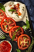 From above of appetizing burrata cheese with sauce and slices of fresh tomato with thyme and basil leaves served on cast iron tray placed on table cloth on wooden table