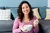 Smiling female with bowl eating breakfast while sitting on sofa in light room looking at camera