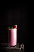 Leckerer gesunder hausgemachter Milchshake mit frischen Erdbeeren, garniert mit grünen Minzblättern, serviert in einem Glas auf einem Holztisch vor schwarzem Hintergrund