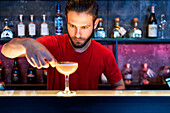 Focused barman decorating cocktail with green leaf served in glass goblet on counter in bar