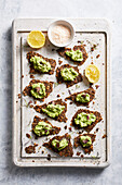 Top view of pieces of crunchy seed crackers with avocado mash served on tray with lemon slices and bowl of grated parmesan in kitchen