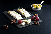 Leckere frische Käsescheiben und reife Weintrauben auf einem schwarzen Brett mit Nüssen und Rosmarinzweigen