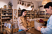 Young female with brown hair in casual clothe standing with small Chihuahua dog in hand in front of woman seller and paying with smartphone in eco friendly shop