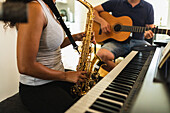Abgeschnittene, nicht erkennbare Frau mit langen lockigen Haaren steht und spielt Saxophon in der Nähe eines positiven Mannes mit Kopfhörern, der in einem hellen Raum sitzt und akustische Gitarre spielt