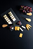 Ansicht von oben auf ein schwarzes Brett mit verschiedenen Käsesorten, reifen Weintrauben und Crackern, die neben Messern auf einer dunklen Fläche liegen
