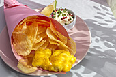 Appetitlich gebratene Pommes frites und Fisch in Papierverpackung auf einem Teller mit Soße auf einem Tisch mit hellem Sonnenlicht in einem Café serviert