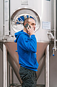 Side view of male entrepreneur talking on cellphone against stainless steel vessels on wet floor in beer factory