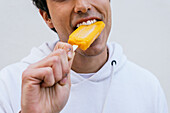 Crop unrecognizable happy male eating sweet orange ice lolly an enjoying taste on white background