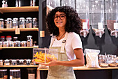 Glad young African American woman in striped apron and glasses looking at camera with smile and putting glass box of dried mango on shelf during work in zero waste grocery store