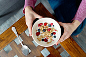 Top view of crop unrecognizable female with bowl of cornflakes and ripe berries with milk for breakfast in light room