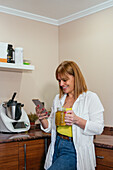 Smiling adult female in casual outfit leaning on counter in kitchen browsing smartphone and holding glass mug of refreshing beverage