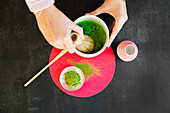 Top view of crop anonymous female with bamboo chasen whipping water and green matcha powder for healthy Japanese drink on bright circle on black table