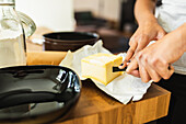 Crop anonymous person cutting fresh butter on wrapping paper into pieces on wooden table with glass jar filled with flour on daytime