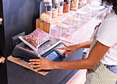 From above side view anonymous young woman pressing buttons on digital balance while weighing plastic bag with chocolate cornflakes during work in eco friendly grocery shop