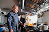 Professional chef doing flambe on food in frying pan with fire while standing near cooker in light kitchen of restaurant