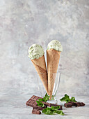 Köstliche Eiskugeln in Waffeltüten mit frischen Minzblättern und Schokolade in einer Glasvase vor grauem Hintergrund