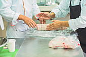 Crop anonymous cooks in uniforms and aprons rolling raw meat in clingfilm while cooking together