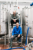 Content male coworkers looking at camera while sitting on stairs between metal tanks in brewing company