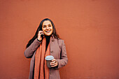 Satisfied female in warm clothes with toothy smile looking away while standing near red wall and having phone conversation
