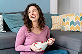 Smiling female with bowl eating breakfast while sitting on sofa in light room looking away