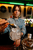 Crop female barkeeper putting ice cube with tongs into wineglass while preparing cocktail in bar with bottles on blurred background