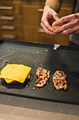 High angle of crop anonymous cook breaking raw egg on grill near cheese and bacon slices while making delicious burger in kitchen
