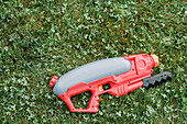Blick von oben auf eine rote und graue Spielzeug-Wasserpistole aus Plastik, die auf einer Wiese im Park liegt