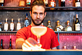 Cheerful barman serving glass with alcohol cocktail on counter in bar and looking at camera