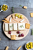 Top view of appetizing cheese served on wooden table with ripe grapes and crackers decorated by rosemary sprigs near olives in bowls on table