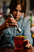 Blurred unrecognizable barkeeper adding lemon zest in glass with alcoholic red old fashioned cocktail while working in bar on blurred background