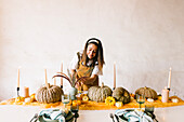 Positive adult Hispanic female designer with long dark hair in apron smiling while decorating table with reed plants pumpkins and candles for Halloween celebration
