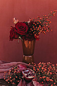 Eleganter Strauß frischer, üppiger roter Rosen und Lilien mit Zweigen roter Beeren neben Stoff und Granatapfel