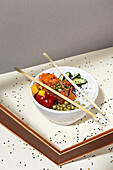 High angle of white bowl with tasty poke dish and chopsticks placed behind frame on table covered with sesame seeds