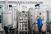 Side view of male coworkers in face masks connecting hose to metal reservoir in beer factory during coronavirus pandemic