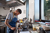 Concentrated young Hispanic male chef with dark hair in casual clothes and apron breaking egg on counter while preparing food during work in restaurant kitchen
