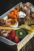 From above brunch box with assorted sliced meats various types of cheese and crispbreads arranged near ripe cup kiwi sweet strawberries and peeled mandarin near jam in glass jar on wooden table near napkin