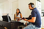 Hispanic Frau mit langen lockigen Haaren stehen und spielen Saxophon in der Nähe von positiven männlichen in Kopfhörer sitzen und spielen akustische Gitarre in hellen Raum in der Tageszeit